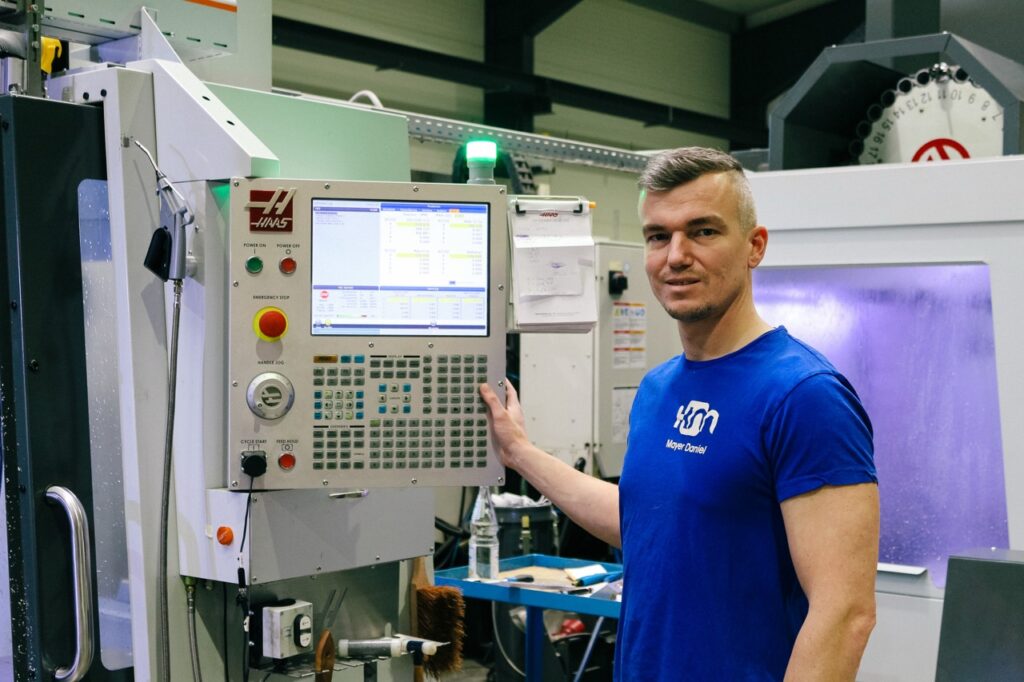 Ein Mitarbeiter steht neben einer Maschine mit vielen Knöpfen und einem Display.